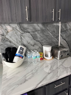 Una encimera de cocina con salpicadero de mármol, una cafetera, un tostador y un bol con toallas de papel y agua embotellada.