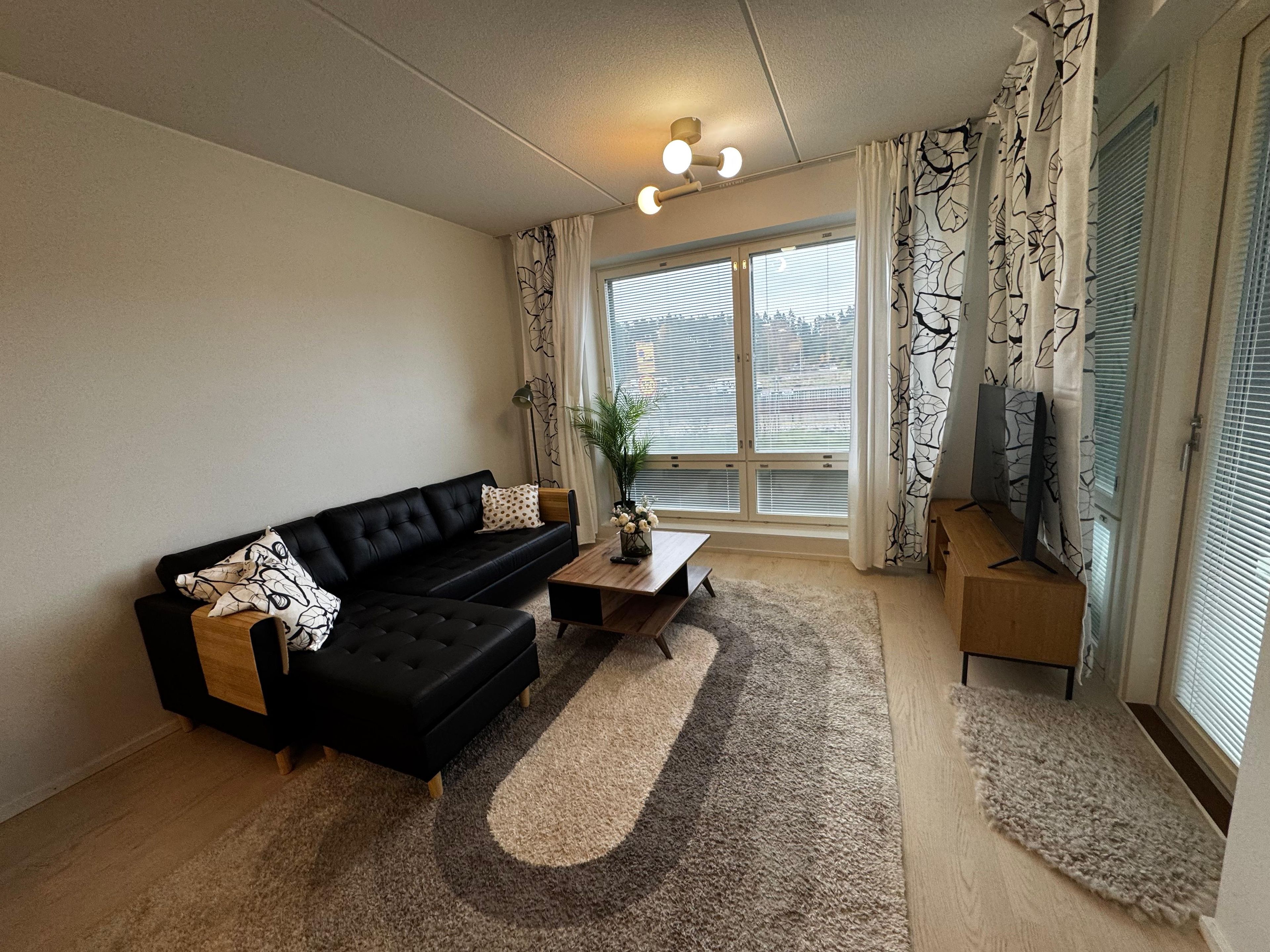 A living room with a black sofa, a wooden coffee table, and a large window with blinds.
