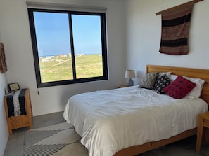 A bedroom with a large window, a bed with patterned pillows, a bedside table with a lamp, and a small wooden bench with a patterned rug. - One bedroom casita with pool and ocean views (Todos Santos)