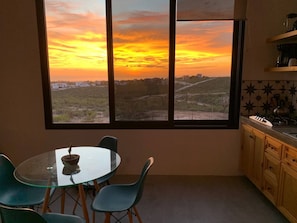 A room with a large window offering a view of a sunset over a landscape, a round glass table with chairs, and a kitchen area with cabinets and a countertop. - One bedroom casita with pool and ocean views (Todos Santos)