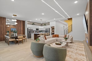 Ein modernes Wohnzimmer mit einer Eckcouch, einem Essbereich mit Tisch und Stühlen sowie einer Küche mit weißen Schränken und Edelstahlgeräten.
