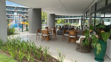An outdoor patio with wooden tables and chairs, a large potted plant, and a view of a playground in the background.