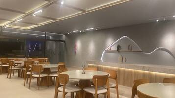 Un comedor moderno con mesas redondas y sillas de madera, una barra con un diseño de iluminación único, y una estantería de pared con objetos decorativos.