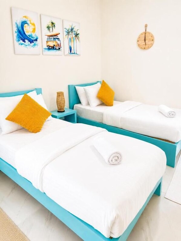 A room with two beds, each with white bedding and a single yellow pillow. There are wall-mounted artworks depicting a wave, a beach scene, and palm trees. A round wooden clock is on the wall. - Ocean Blue Lagoon Maldives (Thulusdhoo Island)