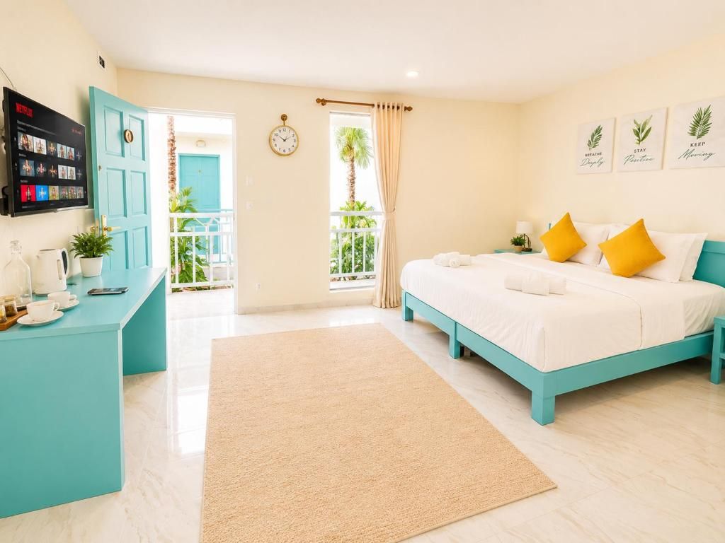 A hotel room with a bed, a desk with a computer, a TV, and a balcony with a view of palm trees.