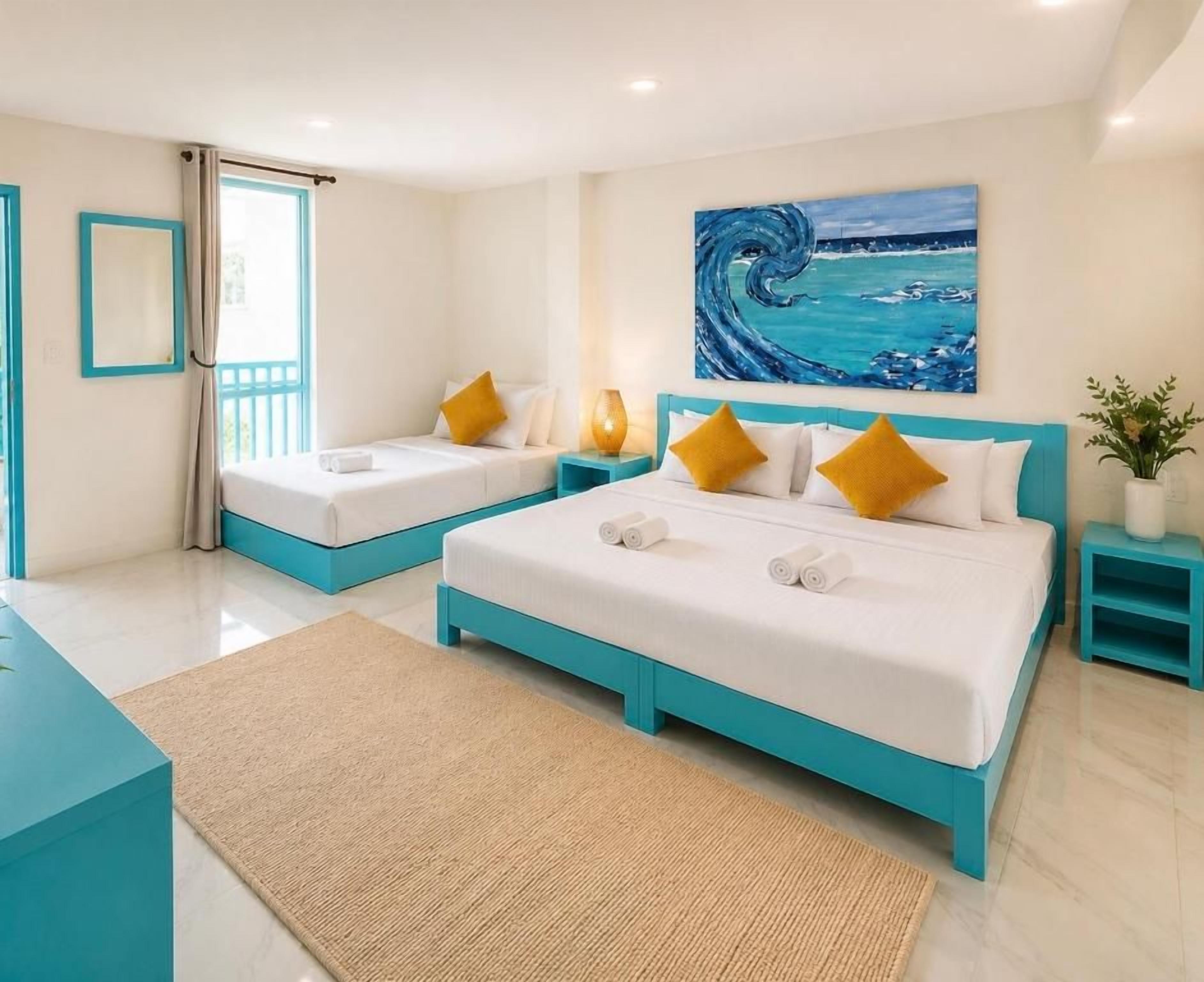 A hotel room with a large bed, two smaller beds, a painting of a wave, and a view of the sea.