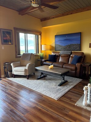 A living room with a brown leather sofa, a beige armchair, a wooden coffee table, and a large window with a view. - Updated 6th -Floor Condo with Stunning Panoramic Grandfather Mountain View’s (Sugar Mountain)