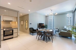 A modern living room with a dining area, a kitchen with an oven, and a large window with blinds. - Sea & Marina View 2BR | Private Beach & Pool (Dubai)