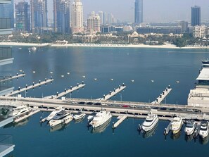 Een jachthaven met diverse boten afgemeerd, een brug naar een stadsgezicht met hoge gebouwen, en een zandstrand op de achtergrond.