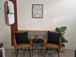Ein Zimmer mit zwei Ledersesseln, einem kleinen Tisch mit roter Vase, einem gerahmten Kunstwerk, einem großen Spiegel und einer Topfpflanze.