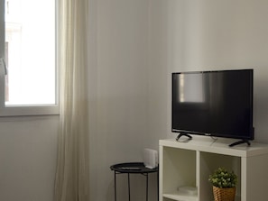 A television on a white stand with a small table beside it, a basket with greenery, and a window with curtains. - Renovated Apartment Near Gran Via (Madrid)