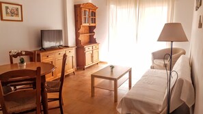 A living room with a sofa, a TV, a wooden cabinet, and a dining area with chairs and a table. - 2B Apartamentos Latin (Nerja)