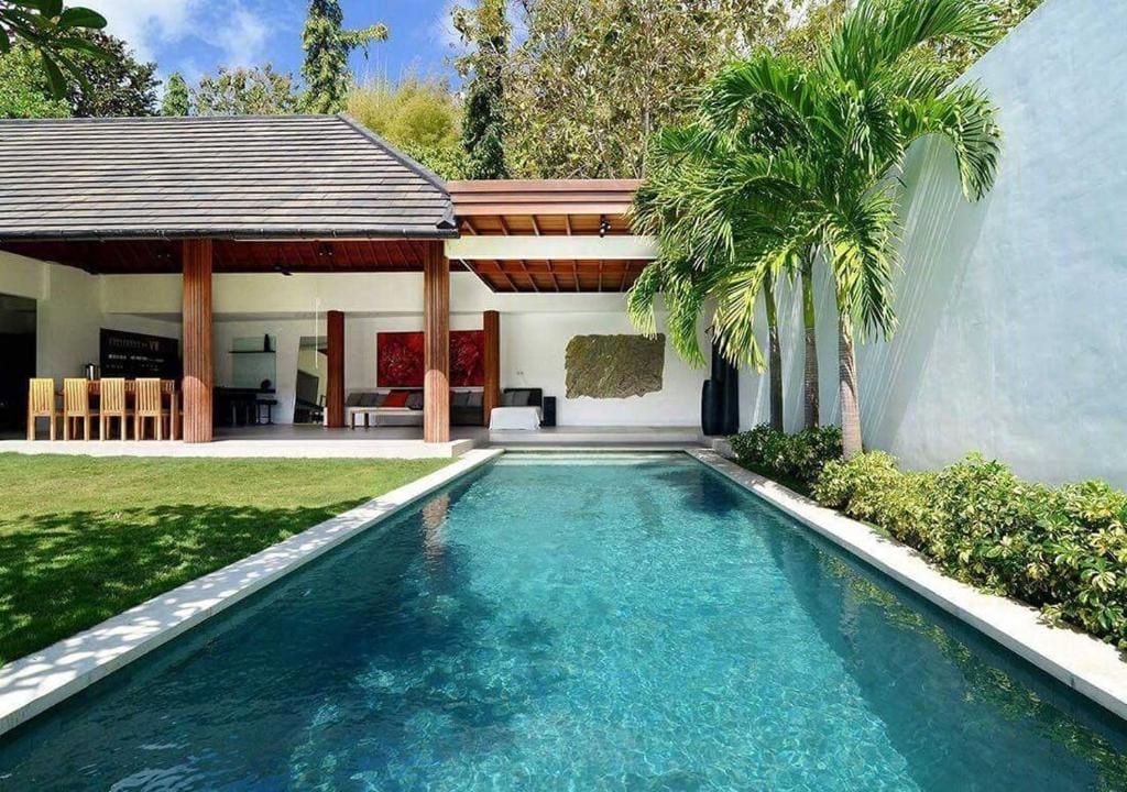 Uma área de piscina moderna com piscina translúcida, rodeada por um deck de madeira e uma área de estar coberta.