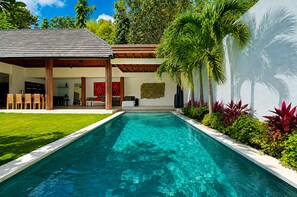Un área de piscina moderna con una piscina azul cristalina, rodeada de abundante follaje y una pérgola de madera.