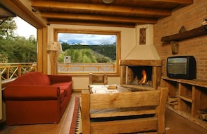 A cozy room with a fireplace, a television, a sofa, and a view of a mountainous landscape through a large window.