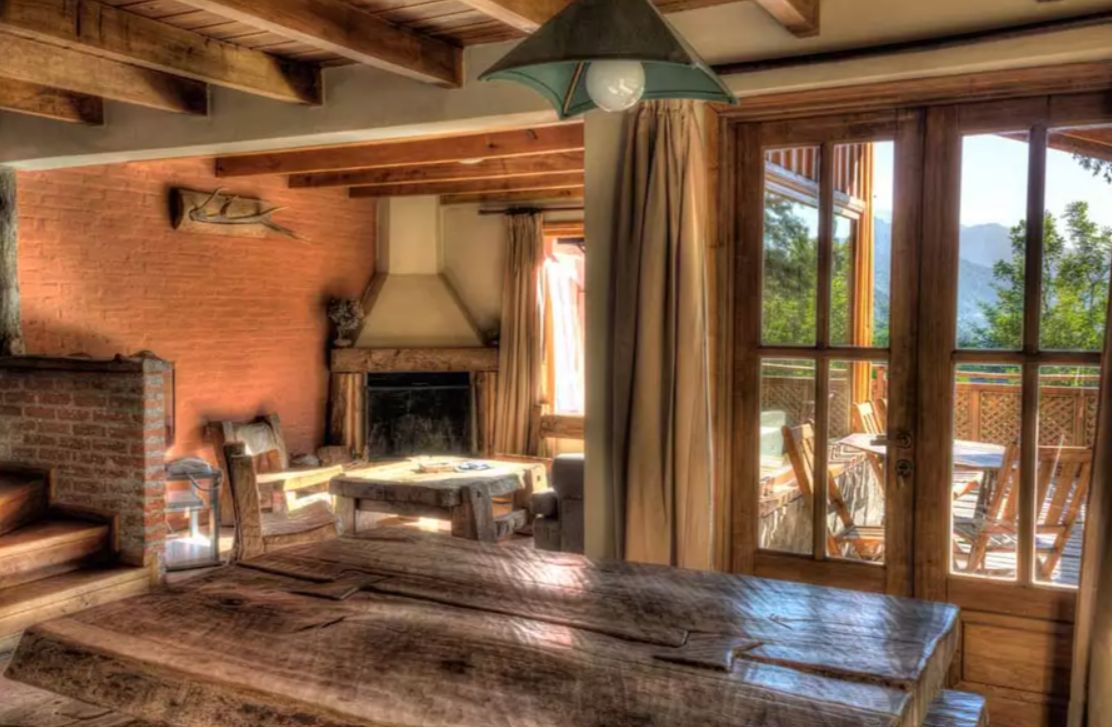 Un interior rústico con pared de ladrillo, vigas de madera, chimenea y un ventanal con vistas a una terraza.