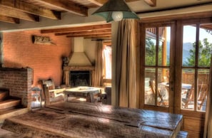 A rustic interior with a brick wall, wooden beams, a fireplace, and a large window overlooking a deck.