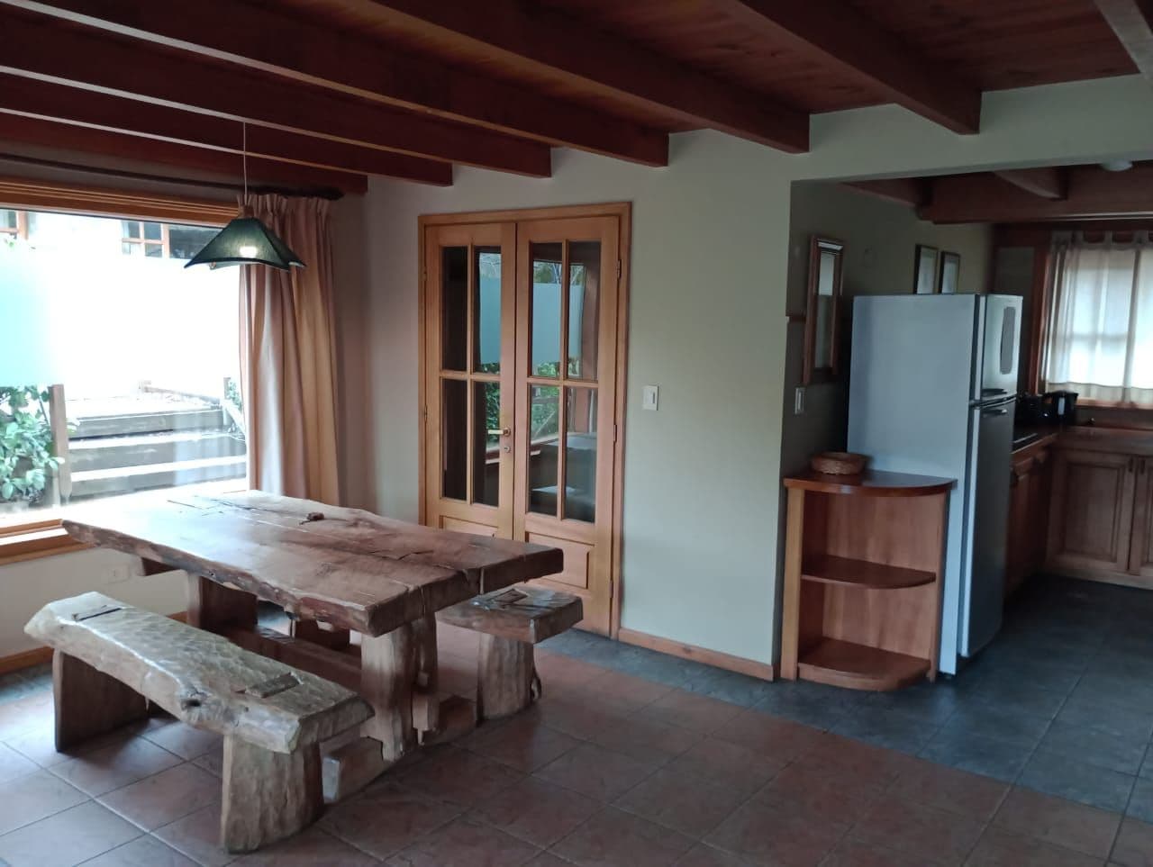 Mesa de jantar de madeira com bancos em um espaço que inclui uma cozinha, armários de madeira e uma janela com cortinas.