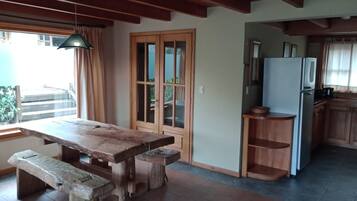 Mesa de jantar de madeira com bancos em um espaço que inclui uma cozinha, armários de madeira e uma janela com cortinas.