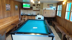 A pool table in a room with wooden walls and benches, a television, and a kitchen area.