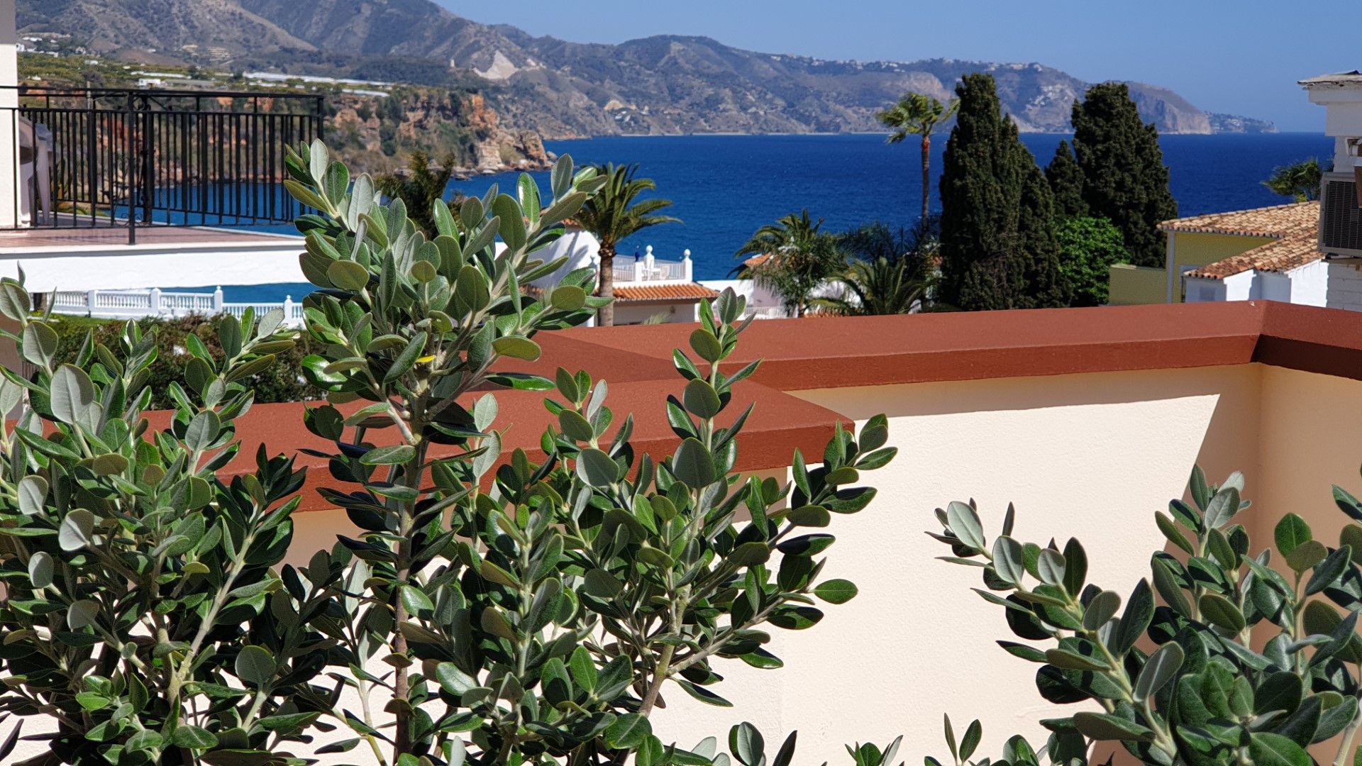 A coastal view with a balcony overlooking the sea, a mountainous backdrop, and Mediterranean vegetation.