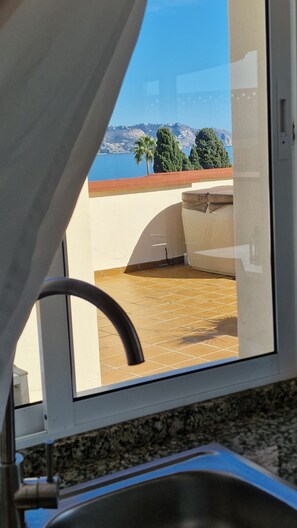 Blick aus einem Fenster mit Metallrahmen, Fliesenboden und Ausblick auf eine Küstenlandschaft mit Bäumen und einem Gewässer.