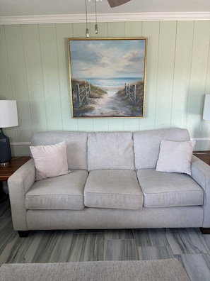 Ein sofas in neutralen Farben mit zwei gemusterten Kissen, ein gerahmtes Landschaftsbild darüber und eine Lampe auf jeder Seite.