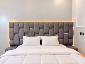 Una cama con cabecera capitonada y dos almohadas, una mesita de noche y un enchufe de pared.