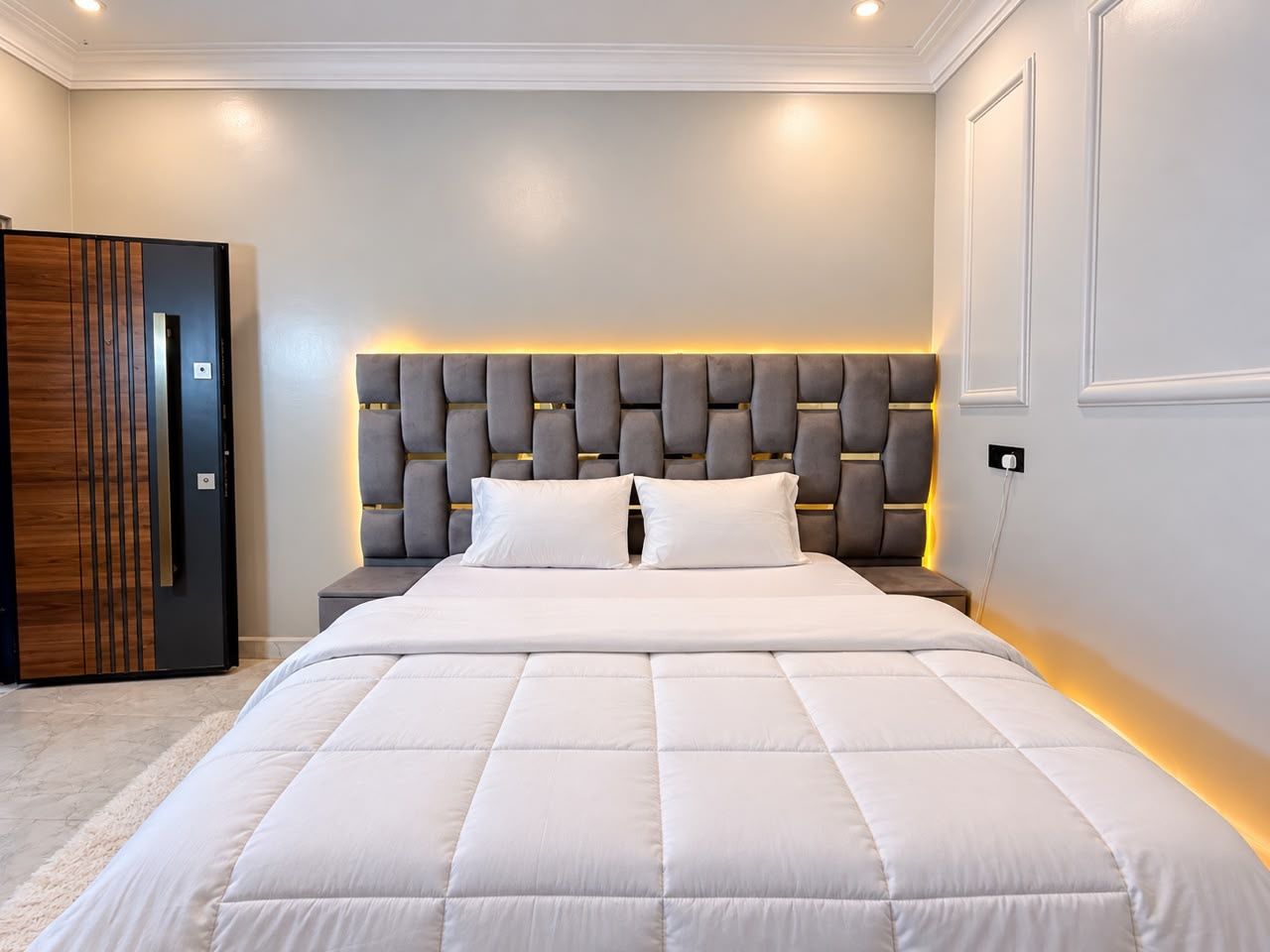 A modern bedroom with a large bed, a headboard with integrated lighting, and a wooden wardrobe.