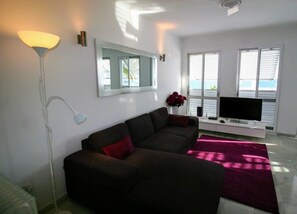 A modern living room with a sectional sofa, a flat-screen TV, a floor lamp, and a large window with blinds. - Carabeo 78X-2 (Nerja)