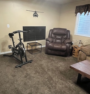 A living room with a stationary bike, a leather recliner, a TV, and a window with blinds. - Newly Renovated Mountain View In-Law Apartment (North Ogden)