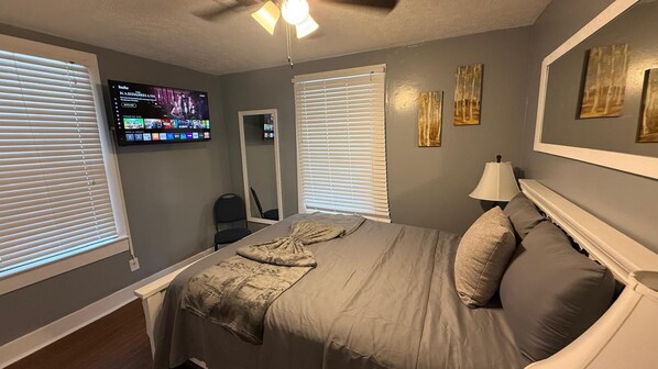 Ein Schlafzimmer mit einem Bett, einem an der Wand befestigten Fernseher, einem Deckenventilator und zwei Fenstern mit Jalousien.