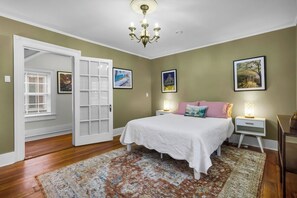 A bedroom with a bed, a nightstand, a chandelier, and a door leading to another room. - Downtown New Bern 3-Bed Home - Yard, Pet-Friendly! (New Bern)