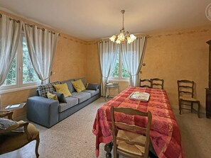 A room with a sofa, a dining table with chairs, a chandelier, and curtains. - Cosy gîte with garden, pets allowed (Commelle-Vernay)
