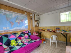 A room with a large world map on the wall, a colorful geometric-patterned sofa, a small white chair, and a shelf with various toys. - Cosy gîte with garden, pets allowed (Commelle-Vernay)