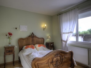 A bedroom with a wooden bed, a bedside table, a nightstand, a window with curtains, and a wall-mounted light fixture. - Cosy gîte with garden, pets allowed (Commelle-Vernay)