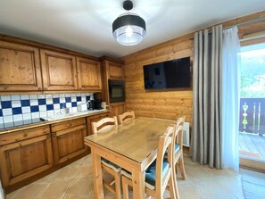 A kitchen with wooden cabinets, a tiled backsplash, a glass-top table, and a large window with curtains. - Renovated apartment, mountain view - 4 guests (Méribel)