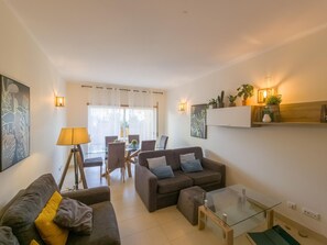 Ein modernes Wohnzimmer mit Sofa, Sessel, Glastisch und einem Essbereich mit Stühlen und Tisch.