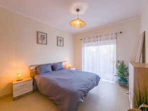 Ein Schlafzimmer mit einem Bett, Nachttischen, einer Hängelampe, einem Fenster mit Vorhängen und einem Blumentopf.
