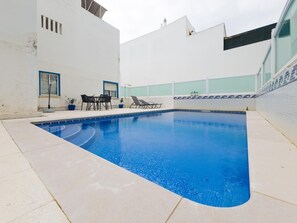 Ein Schwimmbecken mit Fliesenrand, eine Liegebereich mit Stühlen und ein weißes Gebäude mit Fenstern.