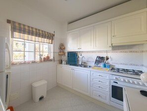 A kitchen with white cabinets, a gas stove, a sink, and a window with a striped valance. - Bright T2 apartment near beaches - 4 guests (Tavira)