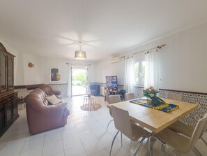 A spacious living room with a sofa, a dining table with chairs, a television, and a large window with curtains. - Villa with air conditioning in Altura - 6 guests (Altura)