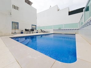 Ein Schwimmbecken mit Fliesenrand, eine Liegebereich mit Stühlen und ein weißes Gebäude mit Fenstern.