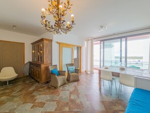 A spacious living area with a large window, a dining table, wicker furniture, and a decorative chandelier. - Eclectic apartment in the heart of Cabanas - 3 guests (Tavira)