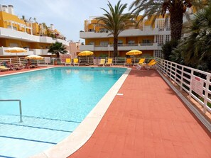 Une piscine entourée d’une clôture blanche et de palmiers, avec des chaises longues et des parasols à proximité.