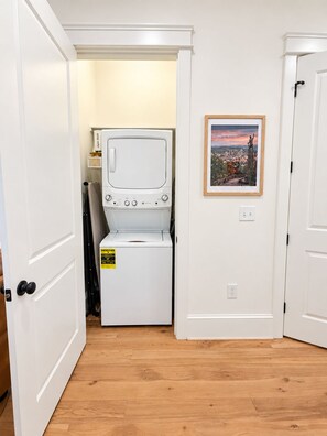 A laundry room with a stacked washer and dryer, a framed cityscape picture on the wall, and wooden flooring. - Luxury 4 Bed 4.5 Bath Townhome-3010 (Homewood)