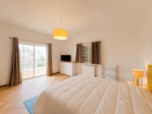 Een slaapkamer met een bed, een televisie, een dressoir en een raam met gordijnen.