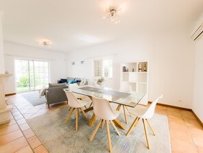 A modern living room with a glass dining table, white chairs, a grey sofa, and a fireplace. - Villa with private pool and AC - Tavira (Tavira)