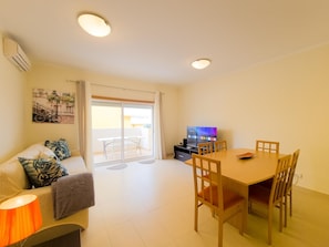 A living room with a sofa, a dining table with chairs, a TV, and a sliding glass door leading to a balcony. - Apartment with AC - 4 guests Near beach (Tavira)