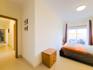 A hotel room with a bed, a small wooden nightstand, a door leading to a bathroom, and a window with curtains. - Apartment with AC - 4 guests Near beach (Tavira)